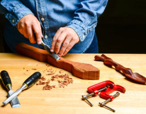 Product photo of a person wearing a denim long sleeved shirt using woodworking tools to whittle a piece of wood on a wooden countertop with a black background.