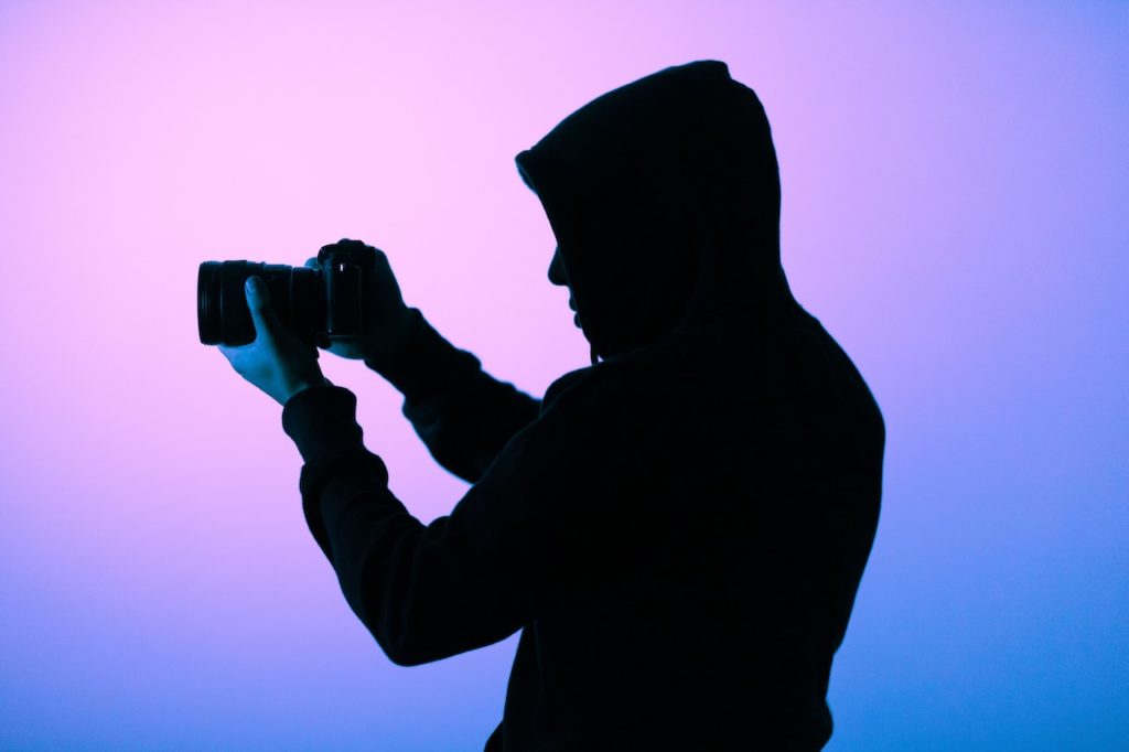 Person holding a camera outstreched in front of them taking a photo. Person has a black hooded sweatshirt on and there are purple and blue gel lights shining in the background.