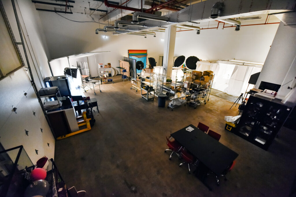 Low lit aerial view of product photography studio displaying lights, tripods, wire racks full of product, computers cameras and a conference table