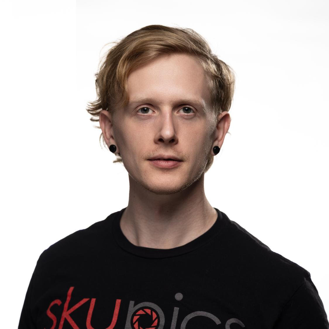 Professional headshot of a man with blue eyes, blond hair, clean shaved wearing a black t-shirt with a red and gray logo Skupics Studios against a white background.