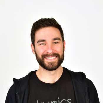 Professional headshot of a man with green eyes, dark hair, dark trimmed beard wearing a black hooded zip up sweatshirt and black tshirt against a white background.